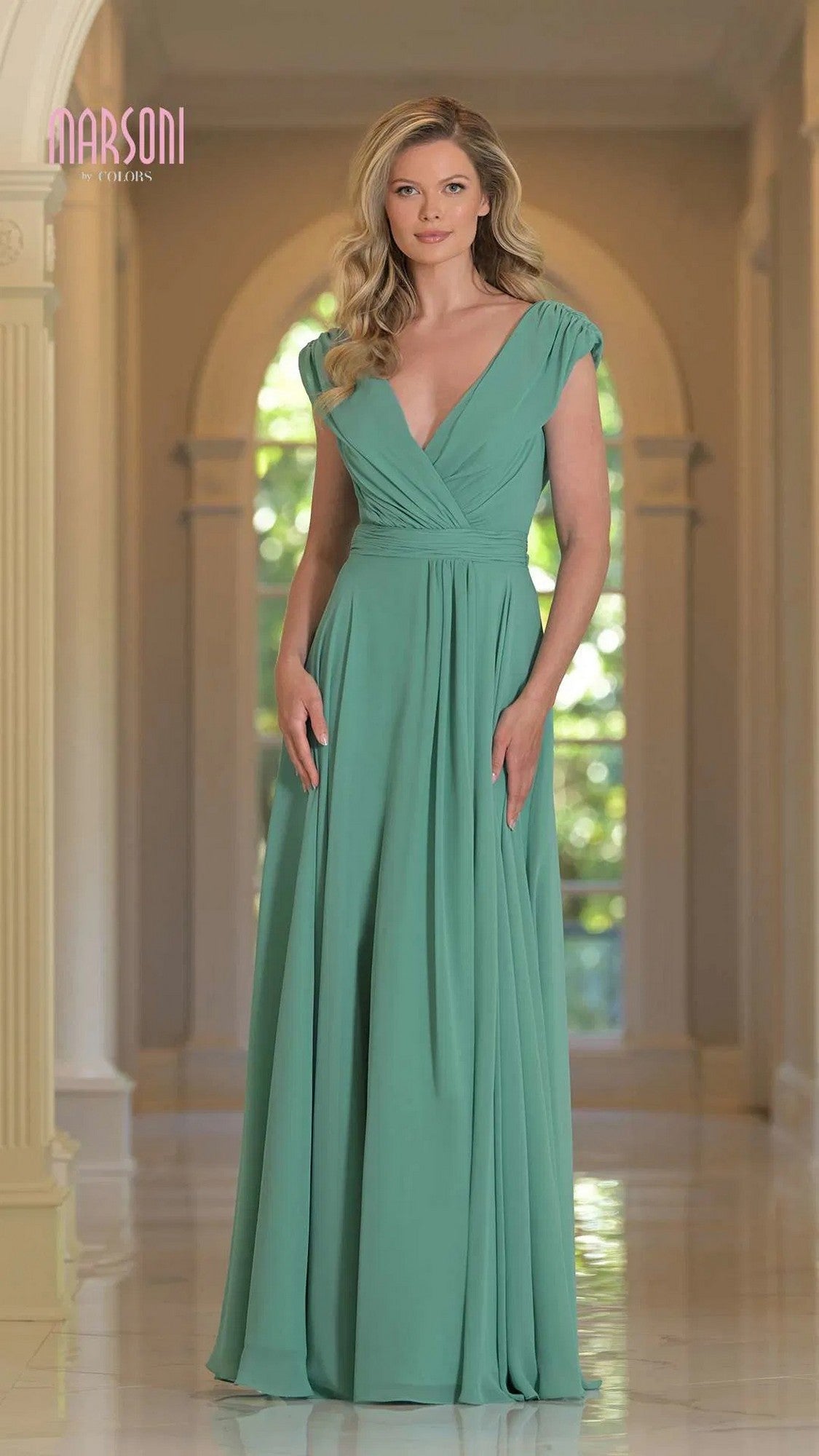 A woman stands indoors in the Marsoni M251, a sage floor-length chiffon formal gown featuring cap sleeves, a V-neckline, and a gathered waist. The softly lit background displays graceful arches.